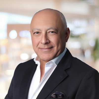 Shows Melih as a middle-aged man wearing a dark blazer and white dress shirt, photographed from the chest up. He is facing the camera with a neutral, approachable expression. The background is softly blurred with warm tones, suggesting an indoor professional setting.