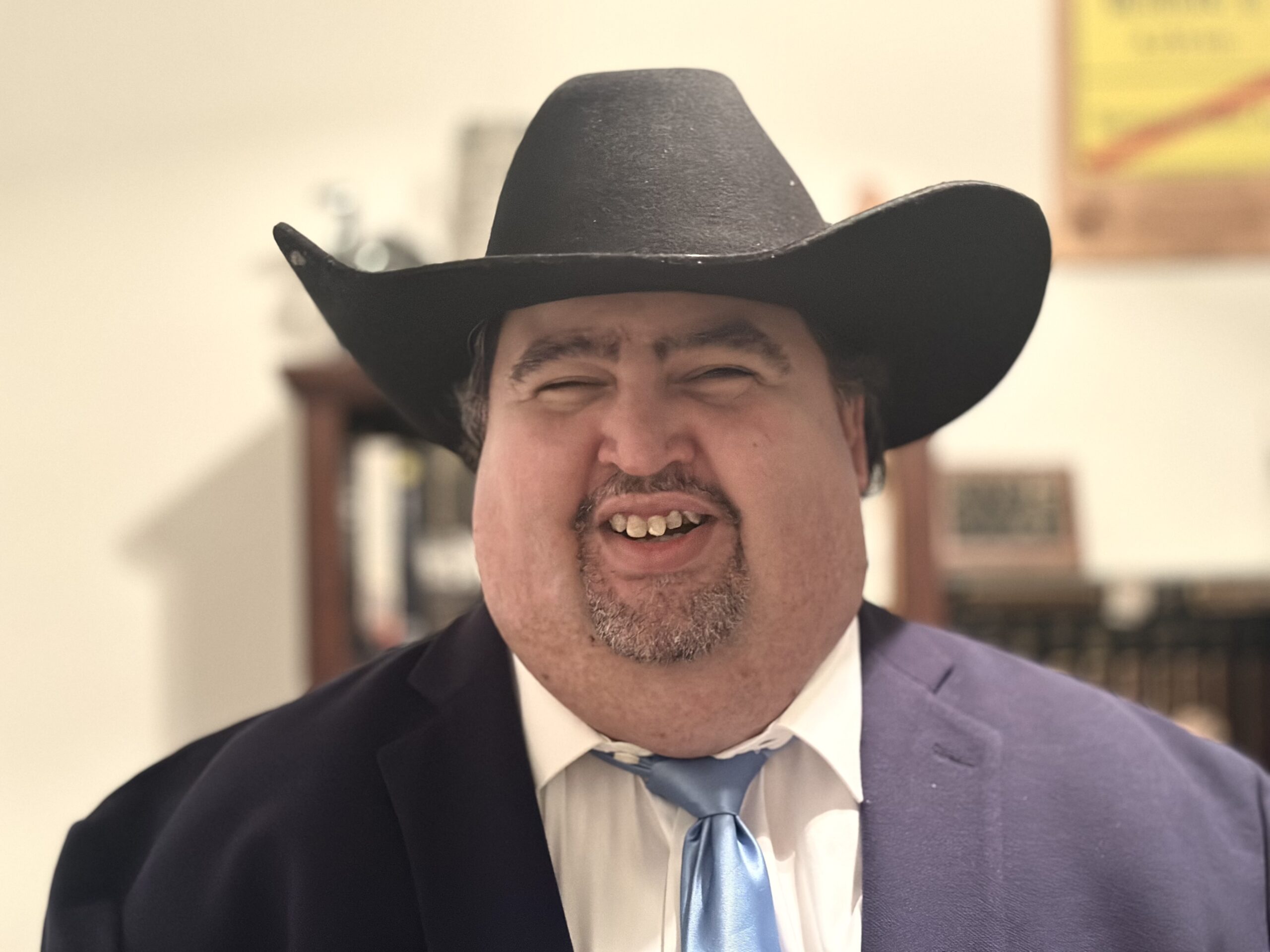Daniel Hodges smiling in a black cowboy hat, navy suit, and light blue tie, standing in front of a bookshelf.