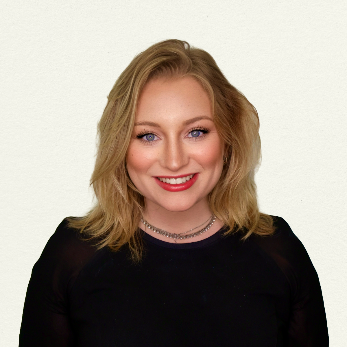 Lauren Harris, a woman with shoulder-length wavy blonde hair and blue eyes, smiling at the camera. She's wearing a black long-sleeve top with a thin silver necklace and red lipstick, against a soft cream background.
