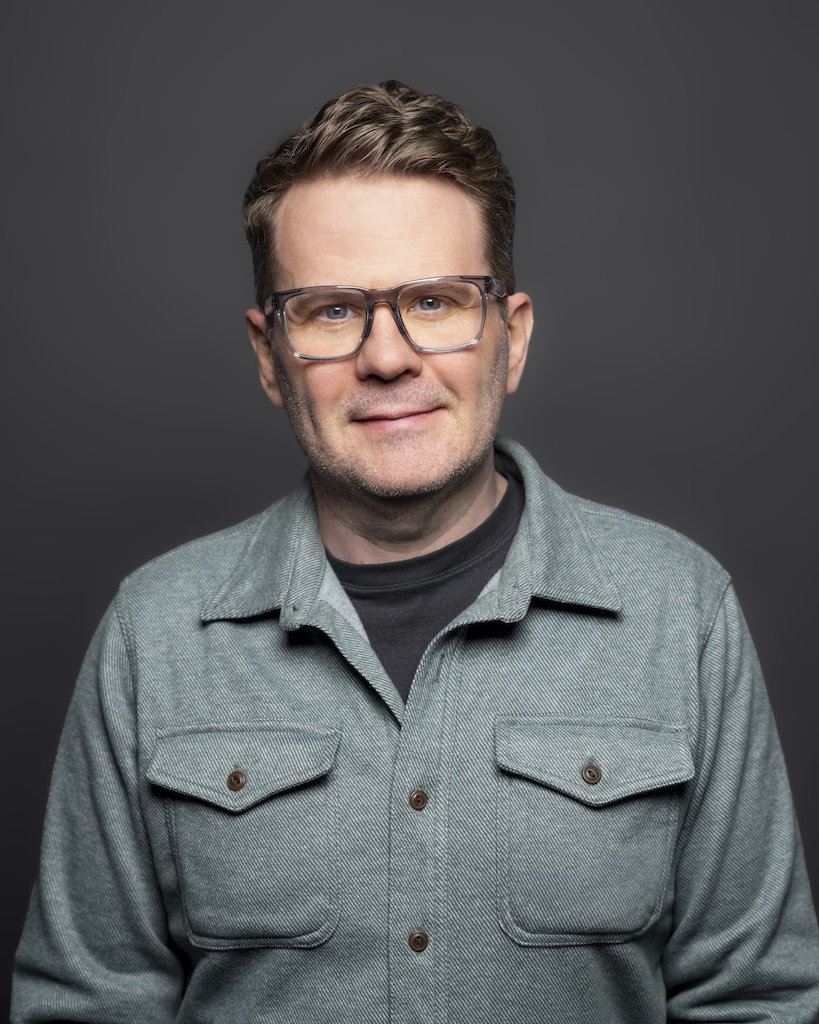 Scott is a smiling man with short brown hair and glasses, wearing a heather gray button-up shirt over a dark t-shirt, photographed against a solid dark gray background.