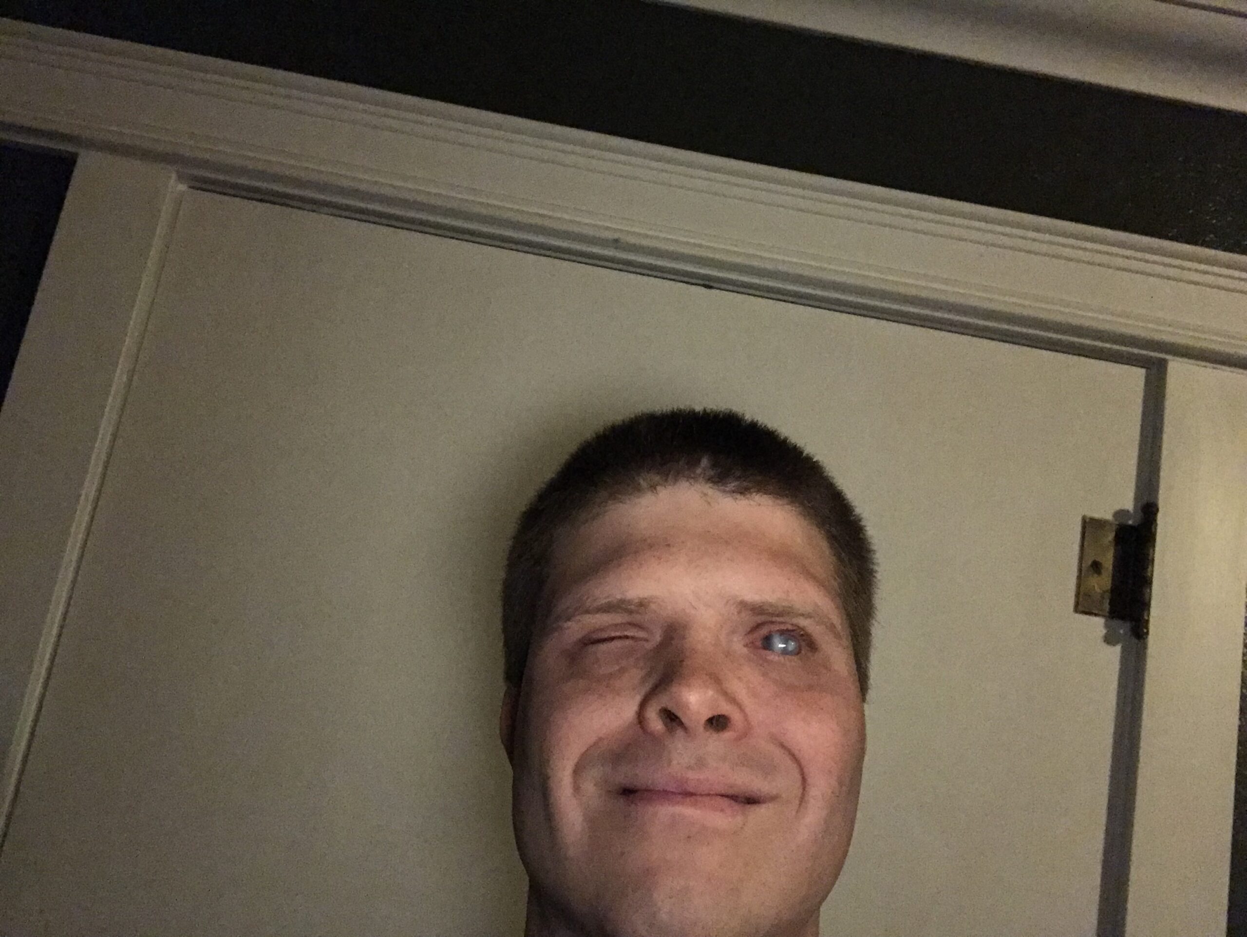 A close-up selfie-style photo of a smiling man with short, closely cropped dark brown hair, taken from a low angle looking slightly upward, standing near a white door.