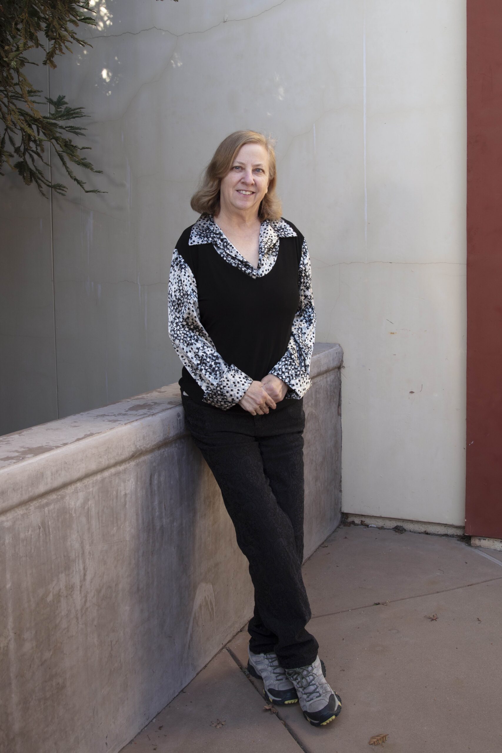A smiling woman with shoulder-length light brown hair, wearing a black and white patterned top and black pants, leaning against a low wall outdoors near a tree branch.