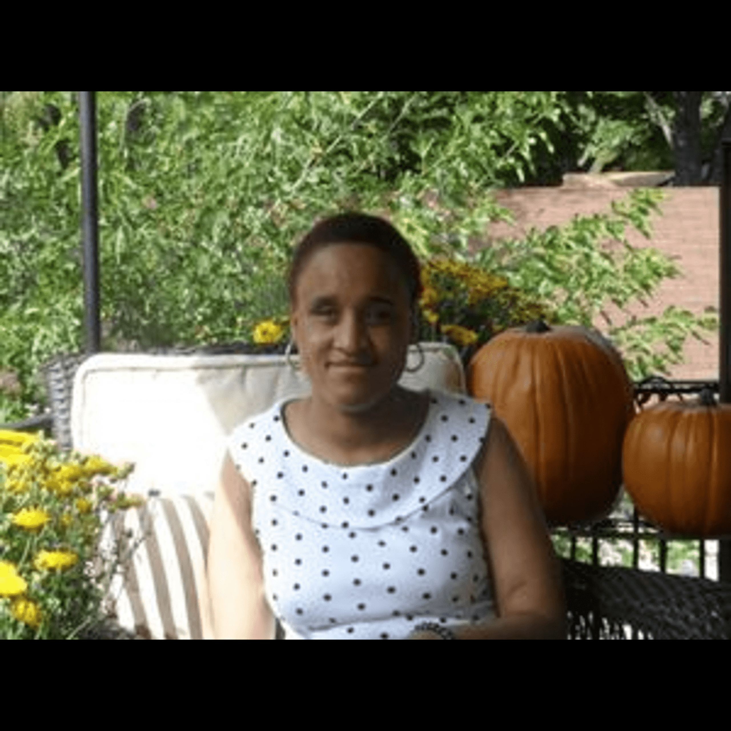 A smiling woman with dark hair pulled back, wearing a white polka dot top, seated outdoors on a porch swing surrounded by greenery, yellow flowers, and decorative pumpkins.