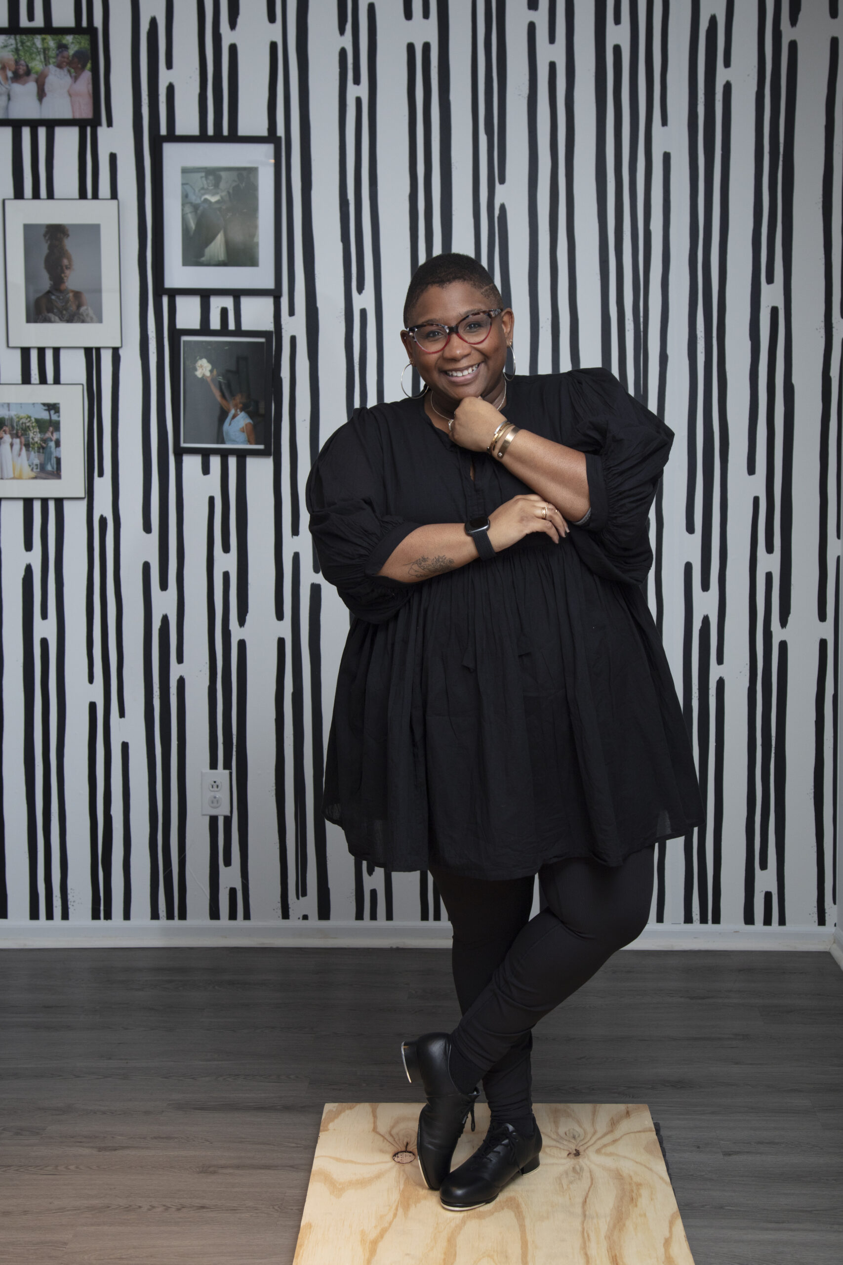 A smiling Black woman with short hair and red-framed glasses, wearing a black dress and hoop earrings, standing on a wooden platform in tap shoes in front of a black-and-white striped wall with framed photos.