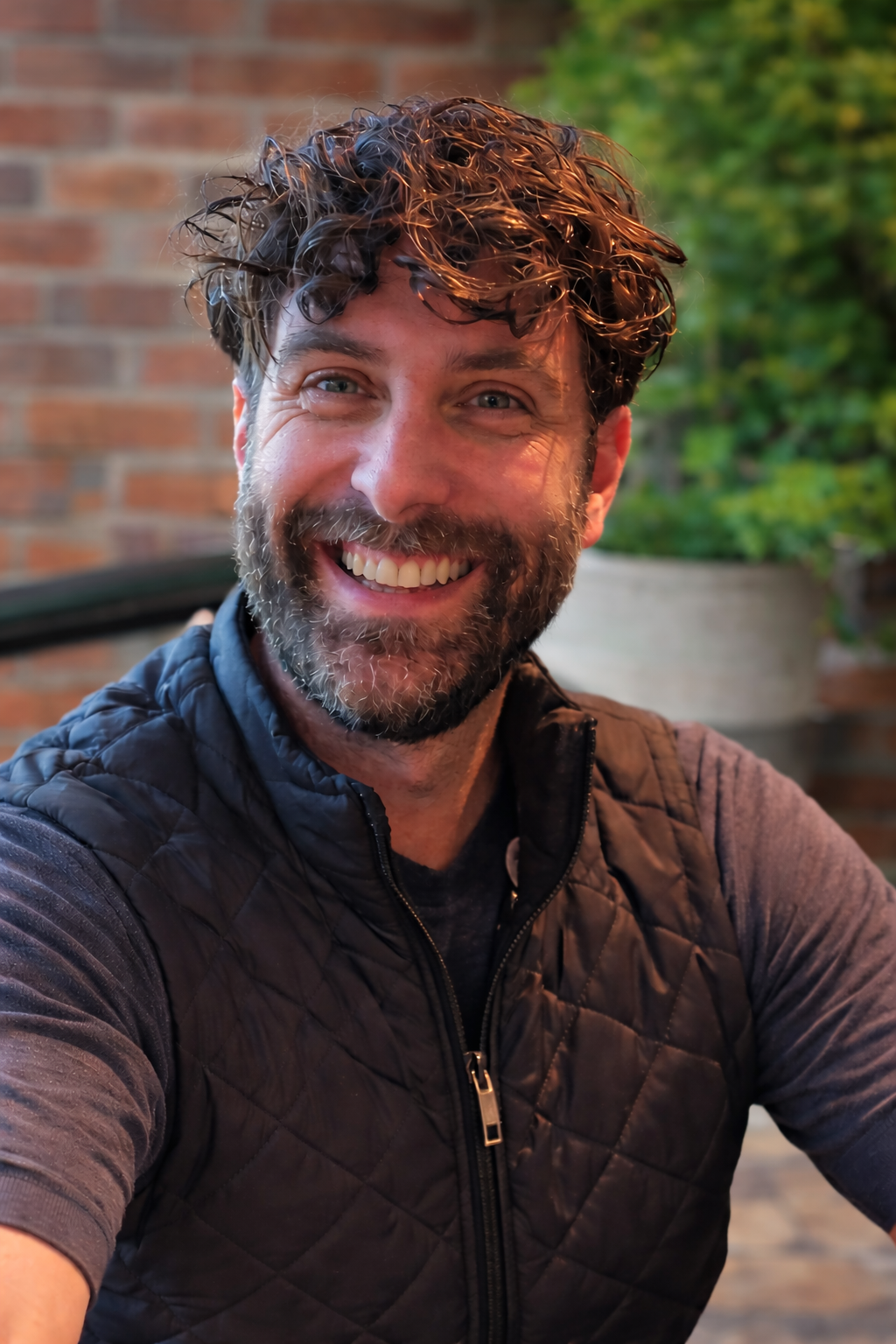 A smiling man with short curly brown hair graying at the temples and a full beard, wearing a dark quilted vest over a gray shirt, photographed outdoors in front of a brick wall and potted greenery.