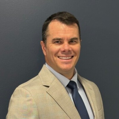 A smiling man with short brown hair, wearing a tan suit, light blue dress shirt, and navy tie, photographed against a dark blue-gray background.